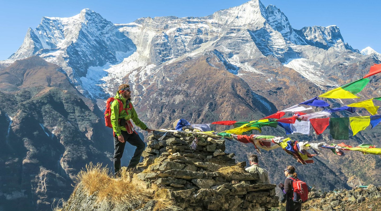 Trekkers on the Everest Base Camp trek by road route near Namche Bazaar with colorful prayer flags and snow-capped Himalayan peaks in the background.