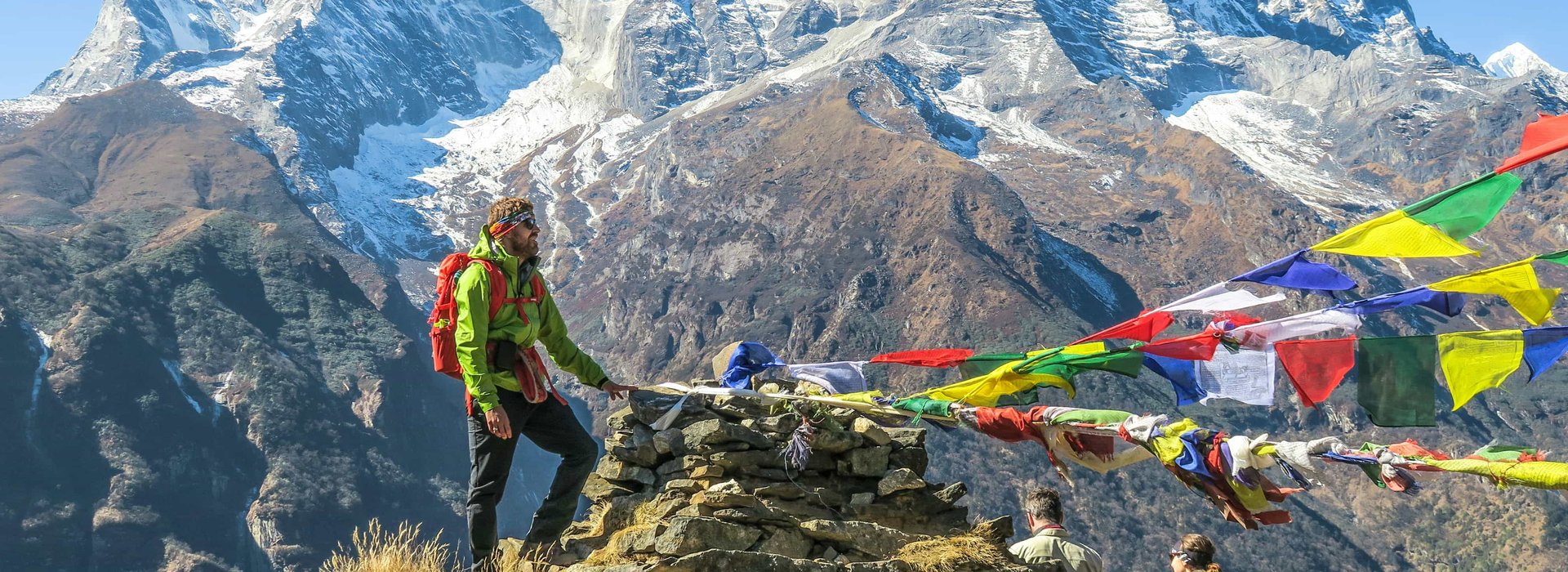 Trekkers on the Everest Base Camp trek by road route near Namche Bazaar with colorful prayer flags and snow-capped Himalayan peaks in the background.