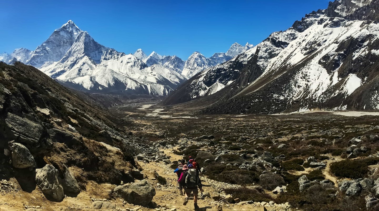 Group of trekkers hiking on rocky trail in the Everest Three Pass trek with Island Peak, surrounded by snow-covered Himalayan mountains under clear blue sky.