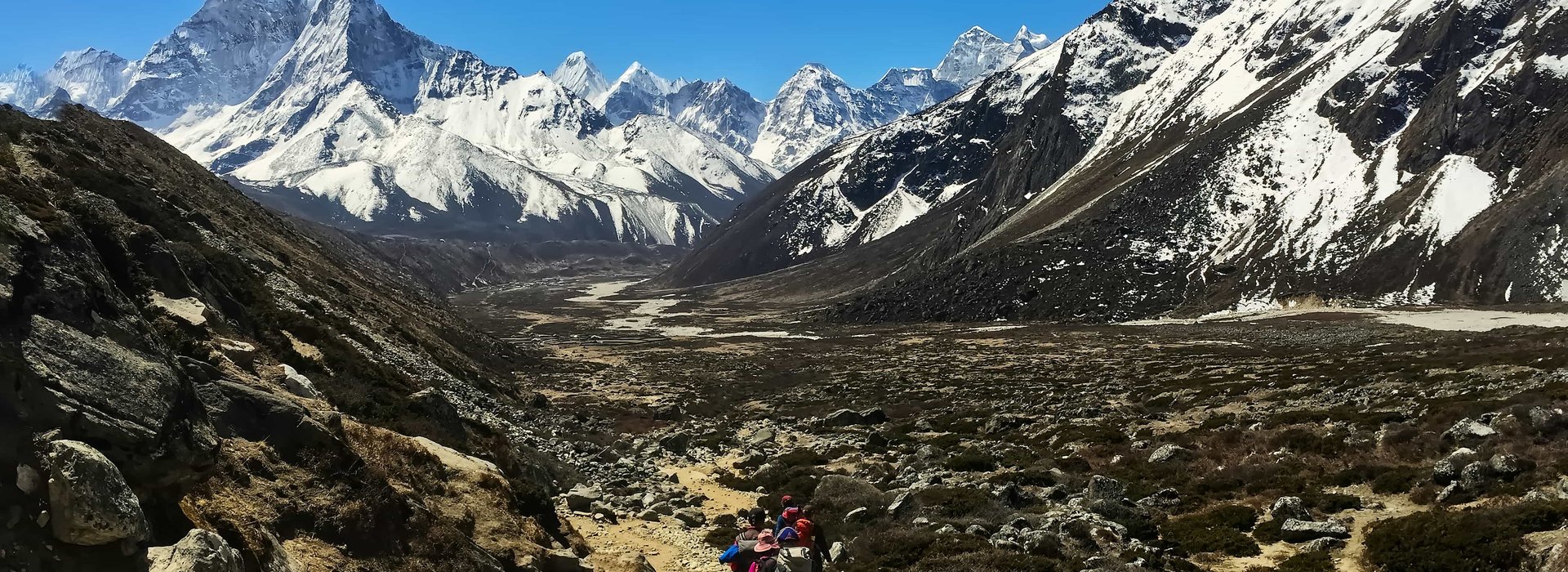 Group of trekkers hiking on rocky trail in the Everest Three Pass trek with Island Peak, surrounded by snow-covered Himalayan mountains under clear blue sky.