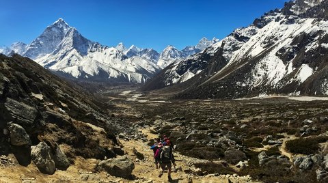 Group of trekkers hiking on rocky trail in the Everest Three Pass trek with Island Peak, surrounded by snow-covered Himalayan mountains under clear blue sky.