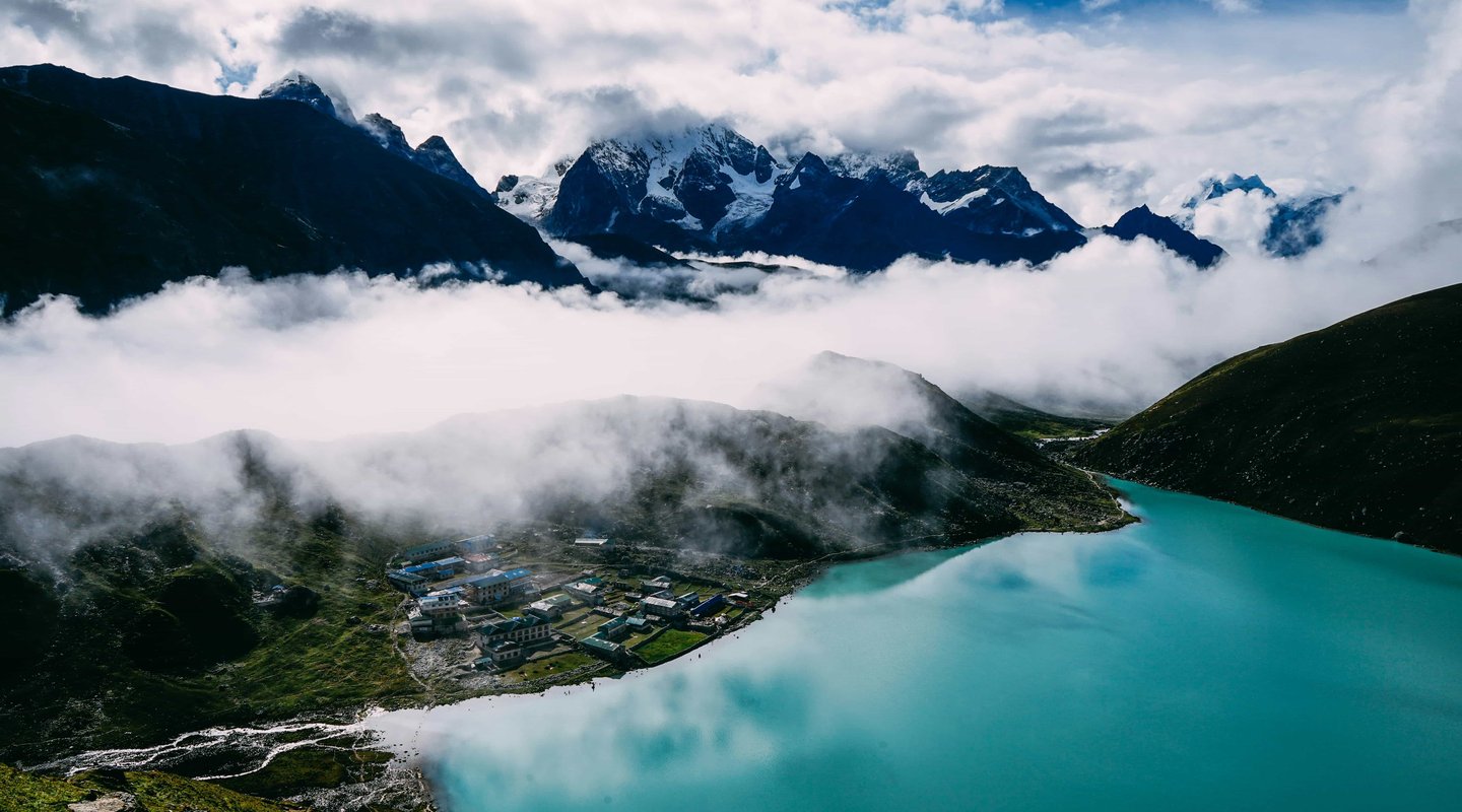Scenic view of Gokyo Lake with turquoise waters, Himalayan peaks, and Gokyo village surrounded by clouds in the Everest region of Nepal.