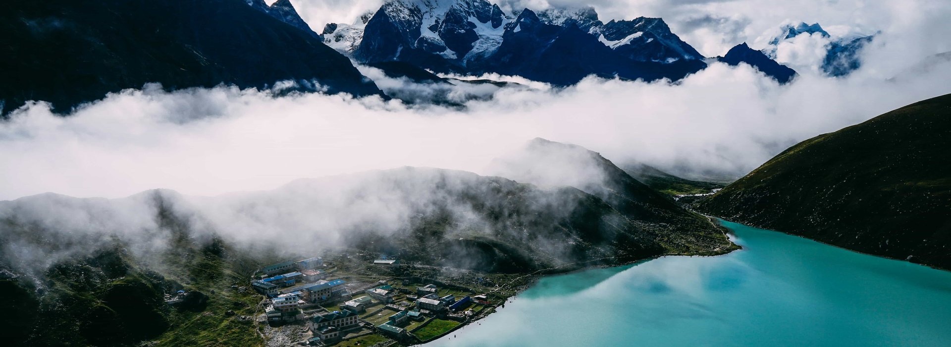 Scenic view of Gokyo Lake with turquoise waters, Himalayan peaks, and Gokyo village surrounded by clouds in the Everest region of Nepal.