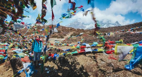 Trekker standing among colorful Tibetan prayer flags on the trail to Everest Base Camp near Jiri, with snow-capped Himalayan peaks in the background