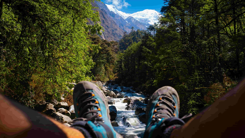 Trekker resting above a mountain river with forested hills and snowy peaks along the Langtang Valley trekking route
