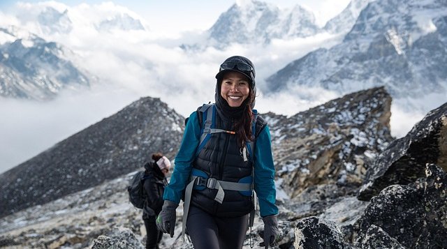 A happy female trekker walking on a rocky alpine trail in the Himalayas, bundled in warm trekking gear with a backpack and gloves, against a dramatic backdrop of snow-capped Himalayan peaks and cloudy skies.