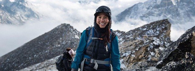 A happy female trekker walking on a rocky alpine trail in the Himalayas, bundled in warm trekking gear with a backpack and gloves, against a dramatic backdrop of snow-capped Himalayan peaks and cloudy skies.