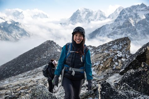 A happy female trekker walking on a rocky alpine trail in the Himalayas, bundled in warm trekking gear with a backpack and gloves, against a dramatic backdrop of snow-capped Himalayan peaks and cloudy skies.