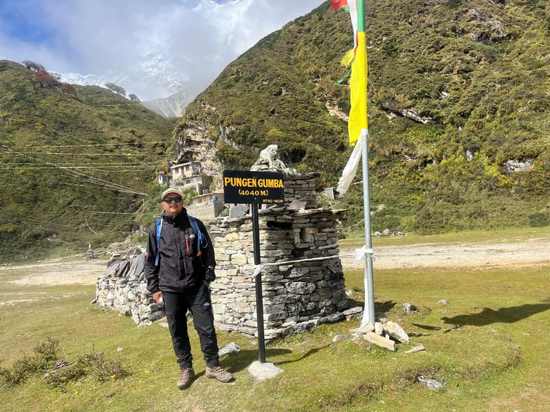 A trekker standing at Pungen Gumba (4040m) on the Manaslu Circuit, with stone chortens, prayer flags, and green mountain slopes in the background under a partly cloudy sky