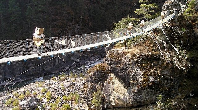 Porters carrying goods across a suspension bridge on the Everest Base Camp trail in Nepal, surrounded by rocky cliffs and pine forests.
