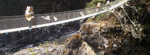 Porters carrying goods across a suspension bridge on the Everest Base Camp trail in Nepal, surrounded by rocky cliffs and pine forests.