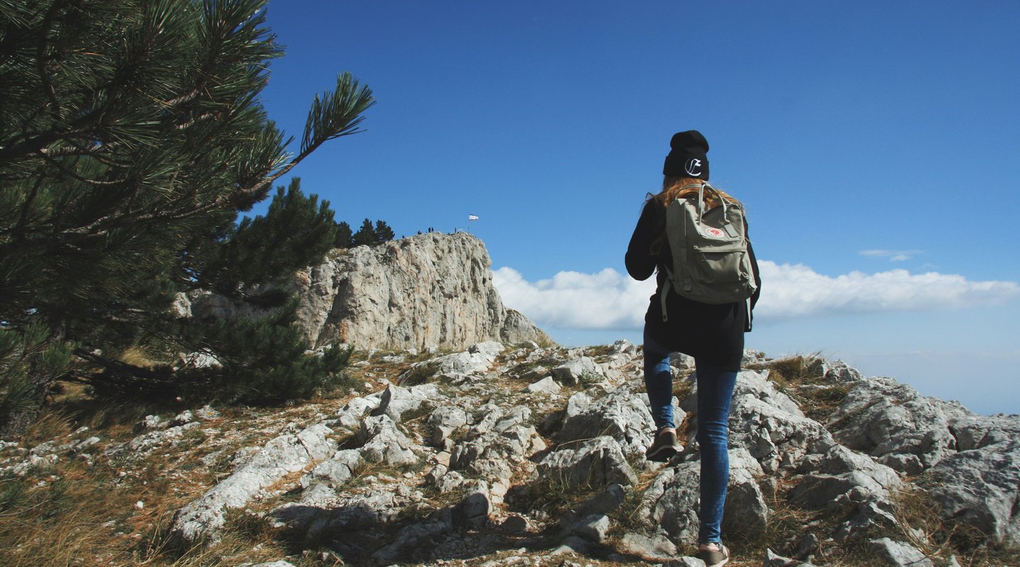 “Woman trekking along a rocky talus path of Manaslu region with snow-capped peaks rising in the distance.”