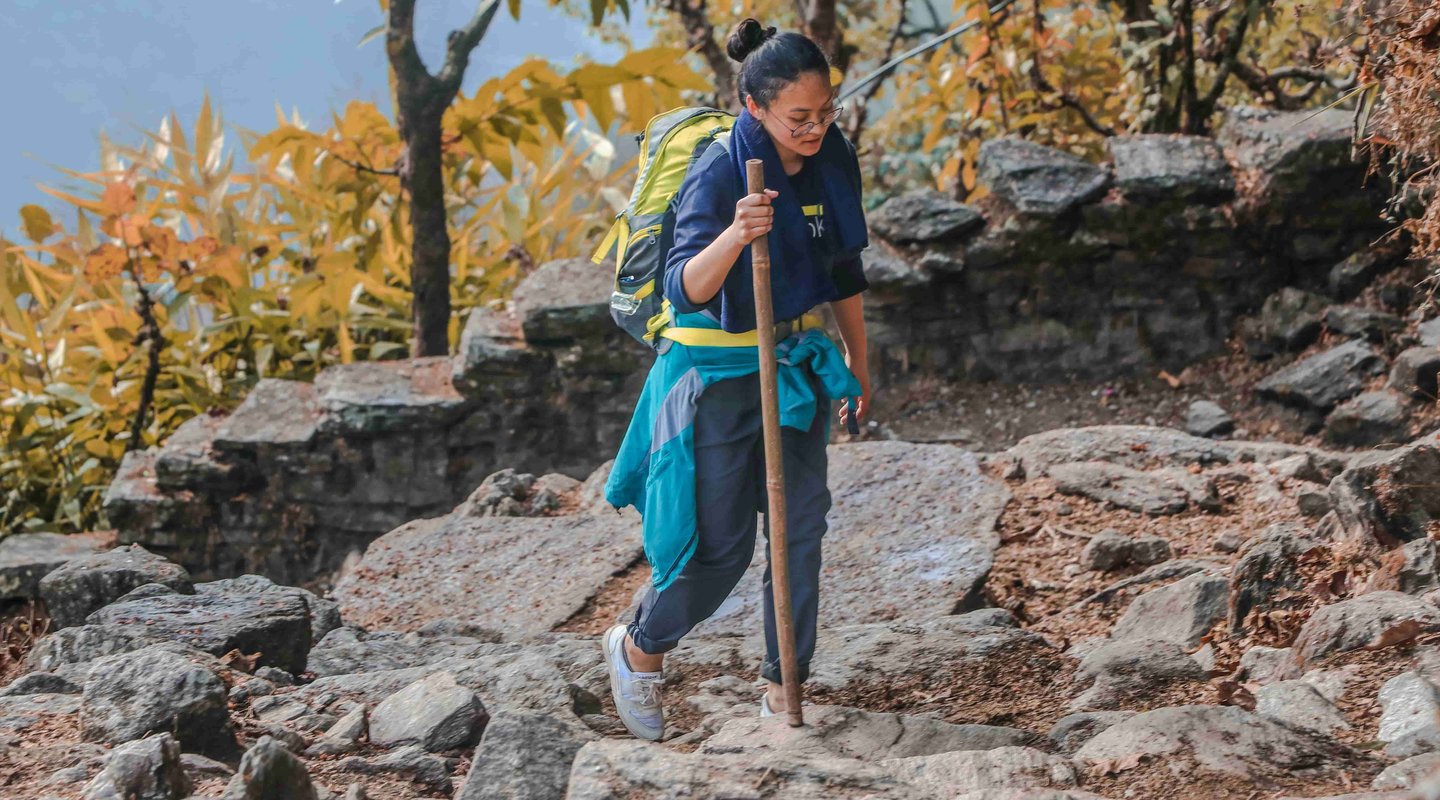 Solo female trekker hiking on a rocky trail in Nepal’s mountains, carrying a backpack and walking stick surrounded by autumn forest scenery — symbolizing safe and empowering solo travel in Nepal.