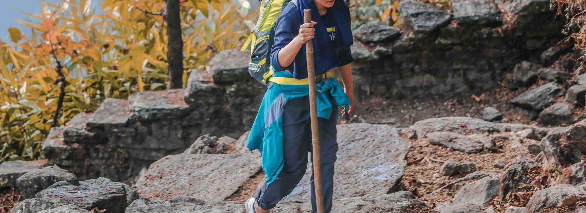 Solo female trekker hiking on a rocky trail in Nepal’s mountains, carrying a backpack and walking stick surrounded by autumn forest scenery — symbolizing safe and empowering solo travel in Nepal.