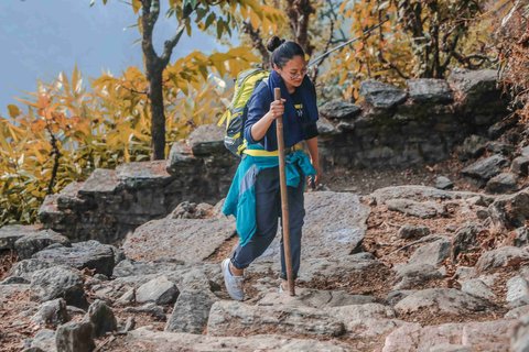 Solo female trekker hiking on a rocky trail in Nepal’s mountains, carrying a backpack and walking stick surrounded by autumn forest scenery — symbolizing safe and empowering solo travel in Nepal.