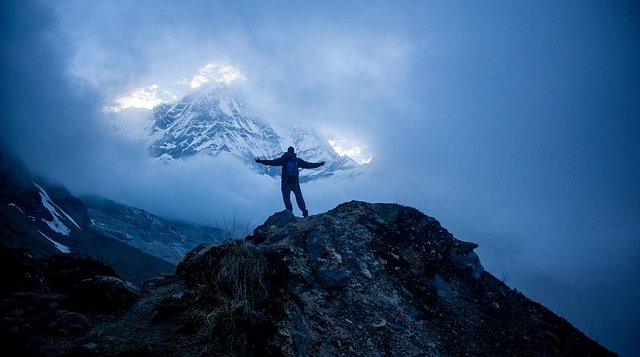 Trekker standing on a rocky ridge surrounded by mist with snow-covered Himalayan peak glowing in the background during spring or autumn trekking in Nepal.