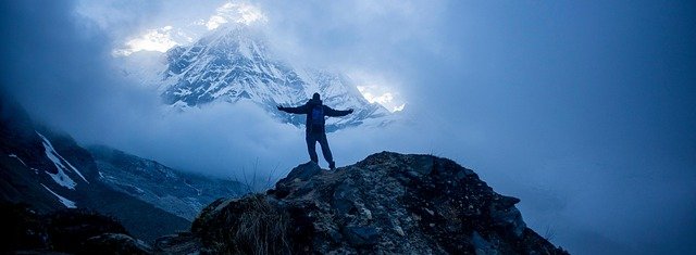 Trekker standing on a rocky ridge surrounded by mist with snow-covered Himalayan peak glowing in the background during spring or autumn trekking in Nepal.