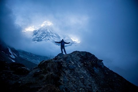 Trekker standing on a rocky ridge surrounded by mist with snow-covered Himalayan peak glowing in the background during spring or autumn trekking in Nepal.