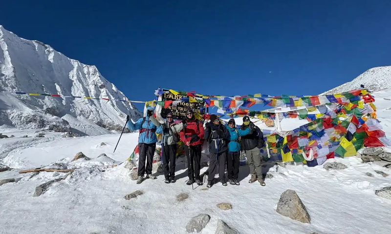 Group of trekkers celebrating at Larkya La Pass on the Manaslu Circuit Trek with colorful prayer flags and snow-covered Himalayan peaks in the background.