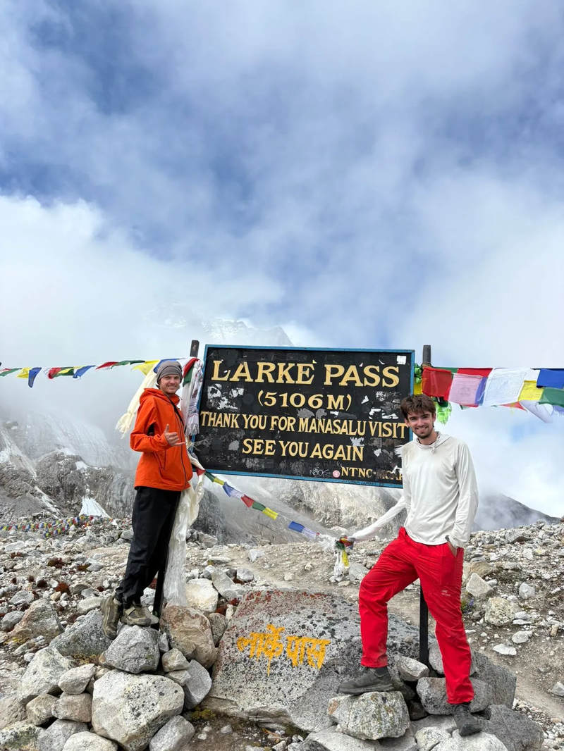 Trekkers from Germany standing beside the Larke Pass signboard during the Manaslu Circuit Trek, surrounded by snow-covered Himalayan peaks.