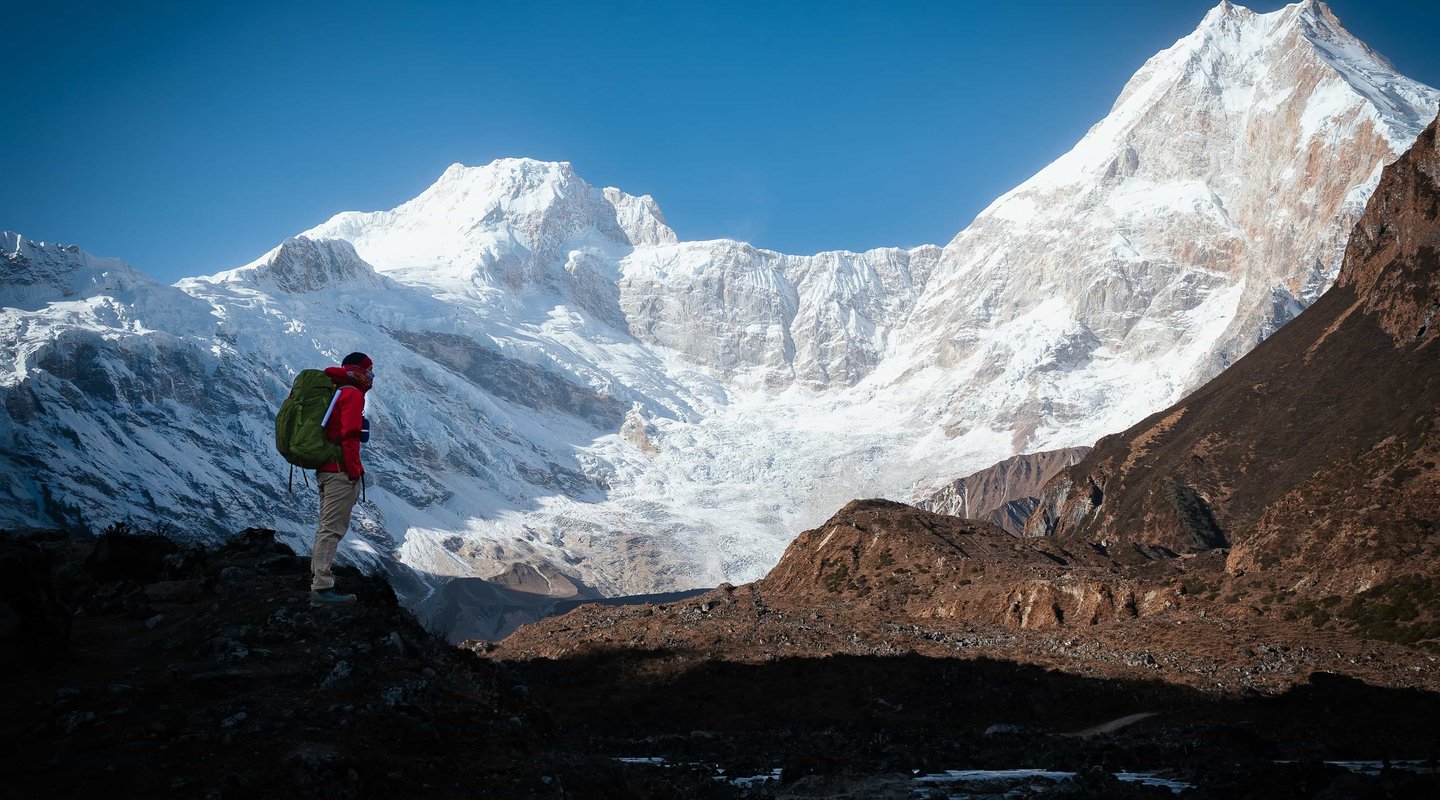 Trekker with backpack standing on a rocky trail overlooking snow-covered peaks of the Himalayas during the Evereat 3 pass trek  in Nepal.