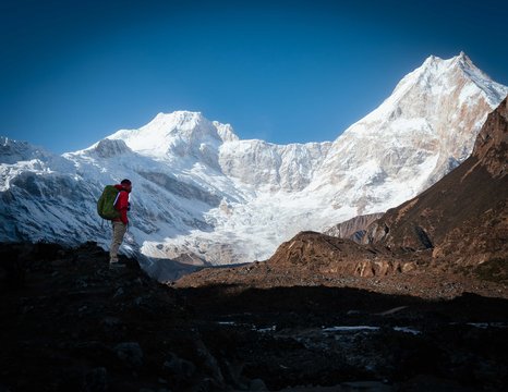 Trekker with backpack standing on a rocky trail overlooking snow-covered peaks of the Himalayas during the Evereat 3 pass trek  in Nepal.