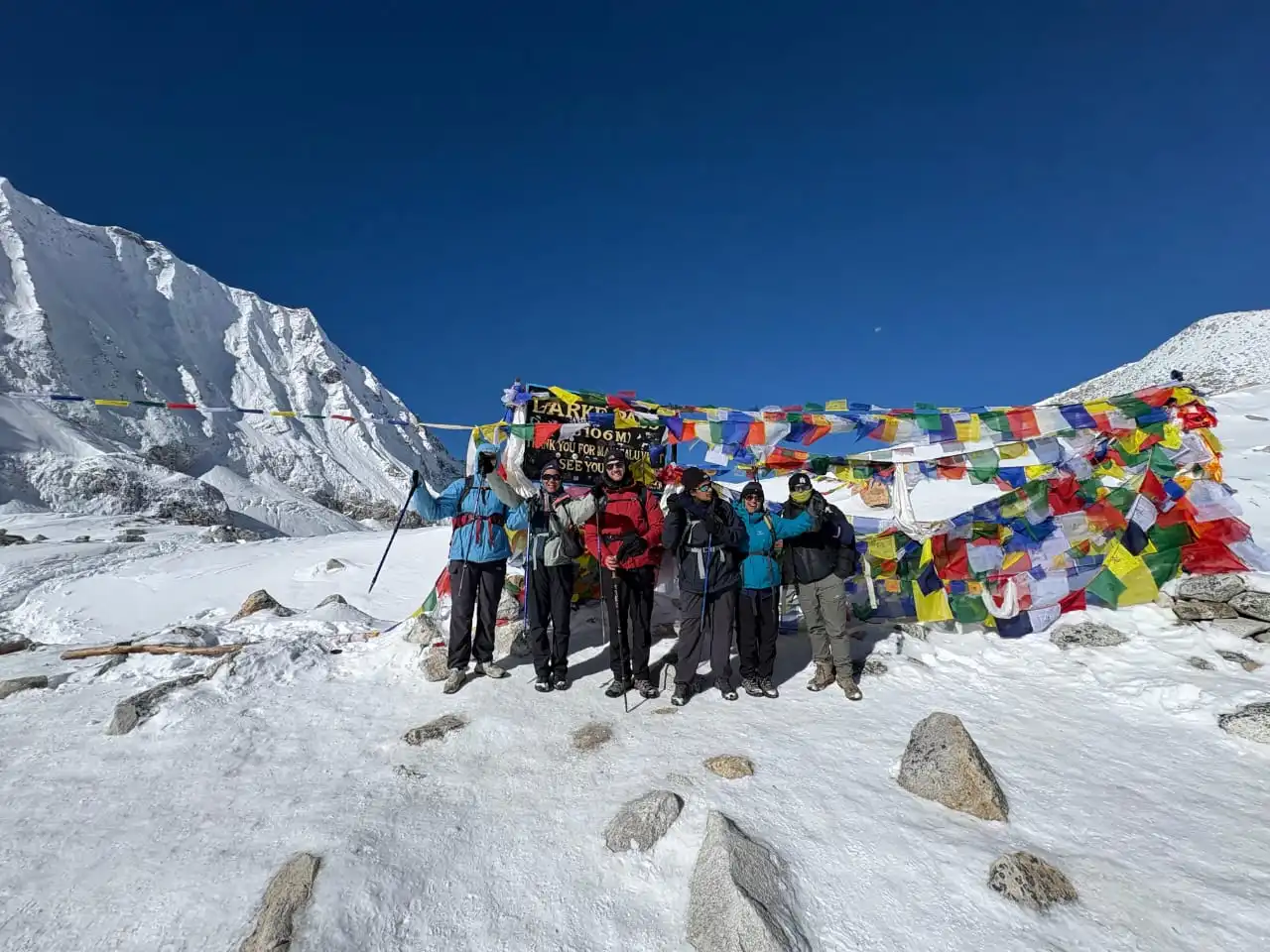 Group of trekkers celebrating at Larkya La Pass on the Manaslu Circuit Trek with colorful prayer flags and snow-covered Himalayan peaks in the background.